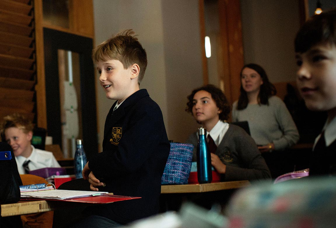 Picture of a boy standing up inside a junior school classroom full of students.