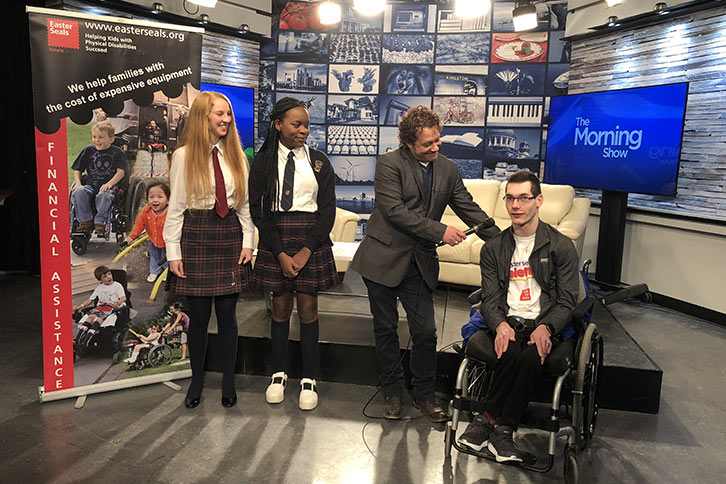 Picture of two students attending a tapping of The Morning Show with the host interviewing a person in a wheelchair