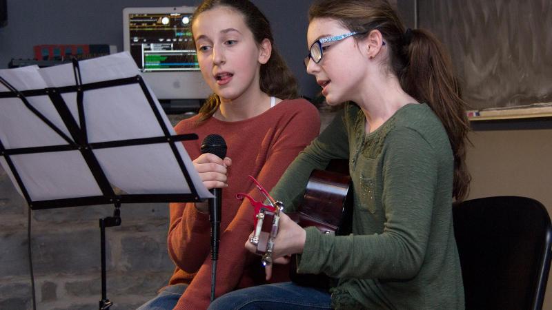 Picture of two students recording music with one student signing and the other student playing the guitar