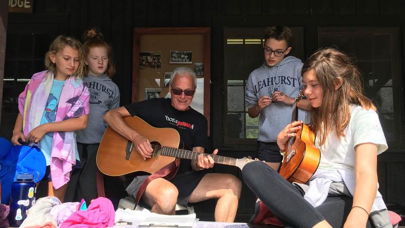 Picture of a teacher playing the guitar for students at a camp activity outdoors
