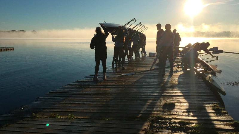 Picture of student holding a row boat over their heads and walking down a deck before putting it in the water