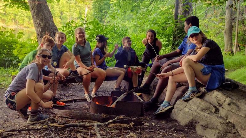 Picture of group of student campers sitting around a camp fire and roasting marshmallows